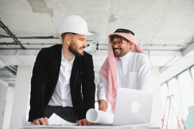 Güvenlik kaskı takan mimar ve Arap yatırımcı bina projesini planlar ve dizüstü bilgisayar kullanarak tartışıyor