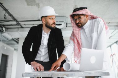 Güvenlik kaskı takan mimar ve Arap yatırımcı planlar ve dizüstü bilgisayar kullanarak gayrimenkul inşaat projesini tartışıyor