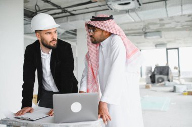 Güvenlik kaskı takan mimar ve Arap yatırımcı dizüstü bilgisayar ve planlar kullanarak gayrimenkul inşaat projesini tartışıyor