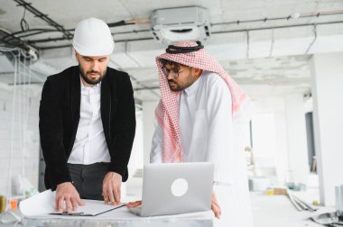 Mimar güvenlik kaskı takıyor ve Arap yatırımcı planları inceliyor ve inşaat sırasında dizüstü bilgisayar kullanıyor.