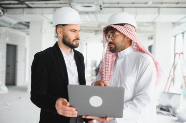 Arap yatırımcı ve inşaat mühendisi bir site ziyareti sırasında bir dizüstü bilgisayarın planlarını inceliyor, bina planlarını tartışıyor ve proje ilerlemesini tartışıyorlar