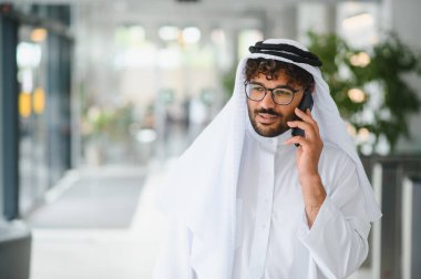 Geleneksel kandura giyen Arap işadamı modern bir ofis binasında telefon görüşmesi yapıyor.