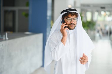 Kendine güvenen Arap işadamı geleneksel kandura giyiyor modern ofis alanında akıllı telefon kullanıyor, kültürel karışımı ve iş iletişimini gösteriyor