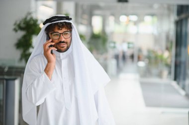 Geleneksel kandura giyen kendine güvenen Arap işadamı modern ofis binasında akıllı telefon kullanıyor.