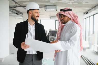 Güvenlik kaskı takan mimar, inşaat halindeki bir binanın içindeki Arap yatırımcının planlarını gösteriyor.