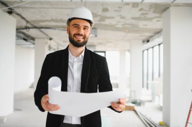 Mimar koruyucu kask takıyor ve elinde planlar tutuyor. Modern binanın inşaat alanını inceliyor.