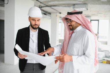 Güvenlik kaskı takan mimar, inşaat halindeki bir binanın içindeki Arap yatırımcının planını gösteriyor.