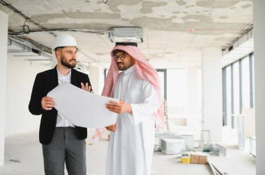 Mimar ve Arap yatırımcı birlikte planları inceliyor, inşaat alanında yapılacak bir gayrimenkul projesinin planlarını tartışıyorlar.