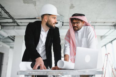 Mimar miğfer takıyor ve yatırımcı geleneksel kıyafetler giyiyor. Bina projesini planlar ve dizüstü bilgisayar kullanarak tartışıyorlar.