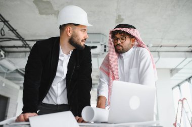 Güvenlik kaskı takan mimar ve Arap yatırımcı bina projesini planlar ve dizüstü bilgisayar kullanarak tartışıyor
