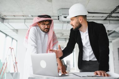 Geleneksel giysiler giyen Arap yatırımcı ve mimar, inşaat halindeki bir binada dizüstü bilgisayardaki bir emlak inşaat projesini tartışıyorlar.