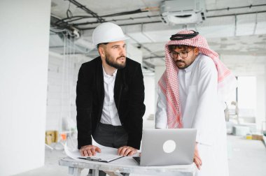 Güvenlik kaskı takan mimar ve geleneksel giysiler giyen yatırımcı inşaat sahasındaki bir ziyarette dizüstü bilgisayardaki bina planlarını analiz ediyor.