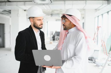 Şapkalı iki mimar bir inşaat alanında dizüstü bilgisayarın planlarını tartışıyorlar.