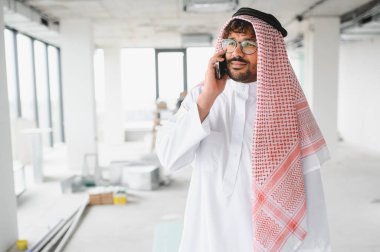 Geleneksel kıyafetler giyen Arap işadamı telefonla konuşuyor, modern iş merkezinin inşaat sahasındaki yatırımları yönetiyor.