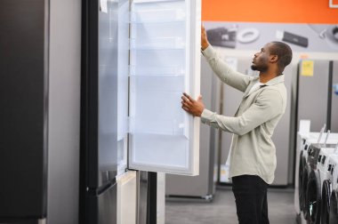 Afrikalı Amerikalı bir adam buzdolabının kapısını açıyor, bir elektronik mağazasında alışveriş yaparken yeni bir buzdolabı seçiyor, seçenekleri ve özellikleri karşılaştırıyor.