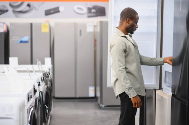 Genç adam, modern bir elektronik mağazasından yeni mutfak aletleri alırken buzdolabı seçeneklerini gözden geçiriyor.