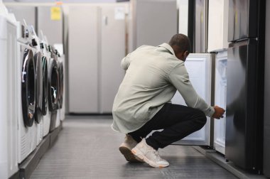 Müşteri çömeliyor, buzdolabının kapısını açıyor, eşyagerdeki özellikleri kontrol ediyor, çamaşır makineleriyle çevrili.