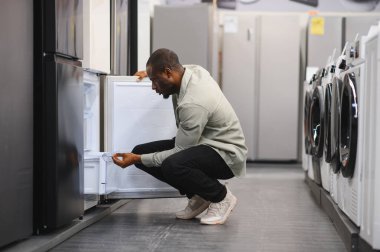 Alış veriş seçeneklerini göz önünde bulundurarak bir mağazada çömelmiş buzdolabını kontrol eden bir adam.