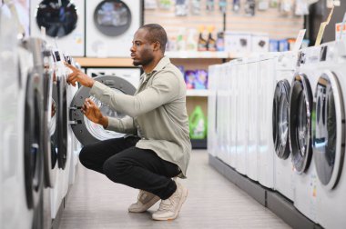 Çömelmiş ve dikkatle yeni bir çamaşır makinesi seçen Afro-Amerikan bir adam ev çamaşırları için modern seçenekleri değerlendiriyor.