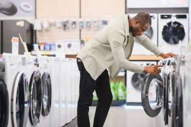 Afro-Amerikalı bir adam, bir alet dükkanından modern bir çamaşır makinesi seçiyor, seçenekleri dikkatlice karşılaştırıyor ve özellikleri inceliyor.