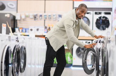Afrika kökenli Amerikalı bir adam modern bir mağazada yeni çamaşır makinelerini karıştırıyor ve satın alma seçeneklerini karşılaştırıyor.