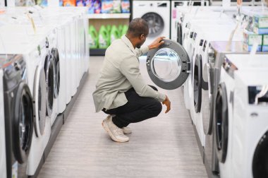 Müşteri bir mağazada alışveriş yaparken çömelmiş çamaşır makinesini inceliyor, ev çamaşırları için seçenekleri değerlendiriyor.