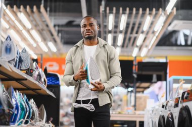 Alet mağazasında yeni bir buhar ütüsü seçen genç adam, alım yapmadan önce özelliklerini ve fiyatlarını karşılaştırıyor.