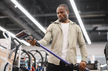 Yeni bir kablosuz elektrik süpürgesi seçmeye odaklanan Afrikalı bir Amerikalı, bir elektronik mağazasının galerisindeki seçenekleri inceliyor.