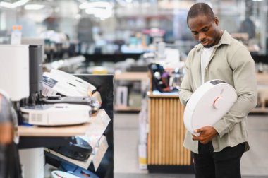 Elektronik mağazasında alışveriş yaparken elinde robot elektrikli süpürge tutan Afrikalı Amerikalı bir adam modern ev temizleme çözümleri seçeneklerini değerlendiriyor.