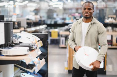 Tüketici elektroniği mağazasından alışveriş yaparken elinde robot elektrikli süpürge tutan Afrikalı Amerikalı bir adam.