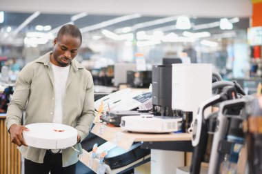 Elektronik mağazasında alışveriş yaparken elinde robot elektrikli süpürge tutan adam ev temizliği için yeni teknolojiler keşfediyor.
