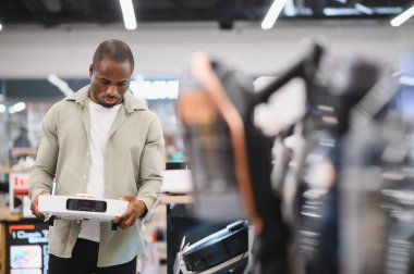 Genç Afrikalı Amerikalı bir adam bir elektronik mağazasında alışveriş yaparken projektör tutuyor, çeşitli seçenekleri araştırıyor ve satın alma kararı veriyor.