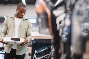 Genç bir adam elektronik mağazasında robot elektrik süpürgesi seçiyor. Satın almadan önce özelliklerini ve tasarımlarını değerlendiriyor.