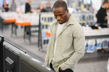 Tüketici elektroniği mağazasında yeni bir televizyon seçen odaklanmış müşteri, yoğun bir perakende ortamında cihazlar için alışveriş yapıyor