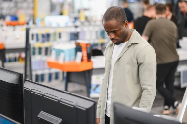 Genç Afrikalı Amerikalı bir adam alışveriş merkezindeki tüketici elektroniği mağazasından alışveriş yaparken televizyonları karşılaştırıyor.