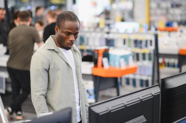 Konsantre olmuş Afro-Amerikan bir adam büyük bir elektronik mağazasında geziniyor, dikkatle yeni bir televizyon alımı seçeneklerini karşılaştırıyor.