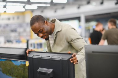 Afro-Amerikalı bir adam alışveriş merkezindeki bir elektronik mağazasında televizyonu dikkatle kontrol ediyor.