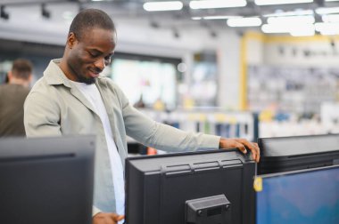 Afrikalı Amerikalı bir adam bir elektronik mağazasından televizyon seçiyor, alışveriş yaparken farklı model ve özellikleri karşılaştırıyor.