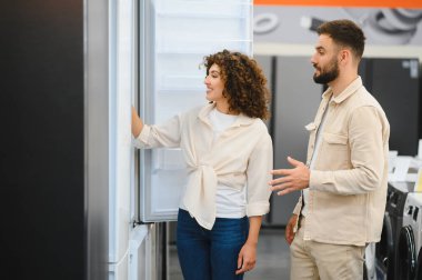 Elektronik mağazasında buzdolabı seçeneklerini inceleyen çift özellikleri tartışıyor ve ev aletleri hakkında karar veriyor.