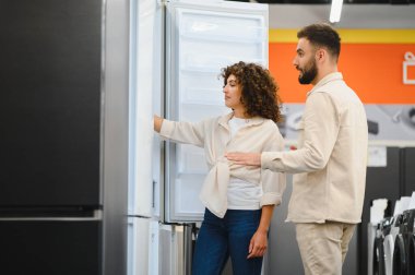 Genç çift elektronik mağazasında yeni buzdolabı seçiyor, özelliklerini inceliyor ve evleri için satın alma seçeneklerini tartışıyor.