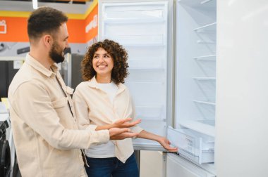 Çift bir elektronik mağazasında buzdolabını inceliyor, özellikleri ve seçenekleri tartışırken evleri için bir şey almayı düşünüyor.
