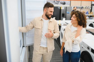 Elektronik mağazasında yeni bir buzdolabı seçerken mutlu çift seçenekleri tartışıyor, satın alma kararları yerine birlikte gülümsüyorlar.