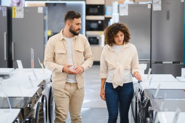 Genç çift yeni bir çamaşır makinesi seçerken seçenekleri tartışıyor. Parlak, modern bir elektronik mağazasında, alışveriş deneyimlerinin tadını çıkarıyorlar.