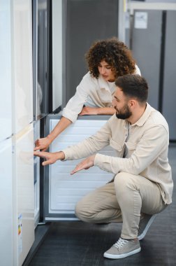 Genç çift bir elektronik mağazasında buzdolabını dikkatle inceliyor, evleri için satın alma kararları alıyorlar.
