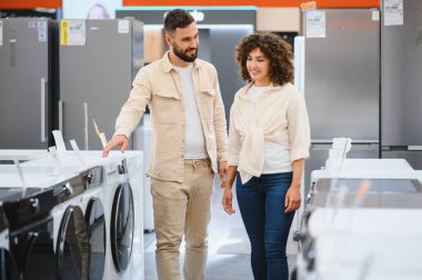 Çift bir elektronik mağazasında ev aletlerini inceliyor, buzdolabı sıralarının arasından mükemmel çamaşır makinesini seçmeye odaklanıyor.