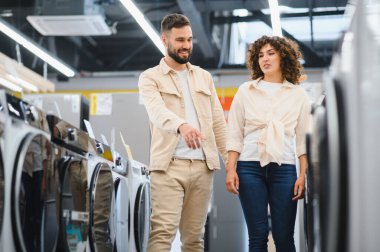 Genç çift elektronik mağazasından çamaşır makinesini seçiyor, adam çamaşır makinelerini işaret ediyor.