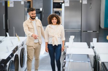 Çift, çamaşır makineleri ve buzdolabı ile çevrili bir elektronik mağazasında ev aletlerini seçmekle meşgul.