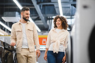Gülümseyen bir çift modern bir elektronik mağazasında ev aletlerini karıştırıyor, seçenekleri tartışıyor ve satın almaları için kararlar alıyorlar.