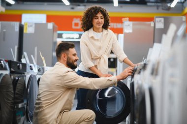 Çift bir elektronik mağazasında ev aletleri seçmekle meşgul, modern bir perakende ortamında çamaşır makineleri ve kurutuculara odaklanıyor.