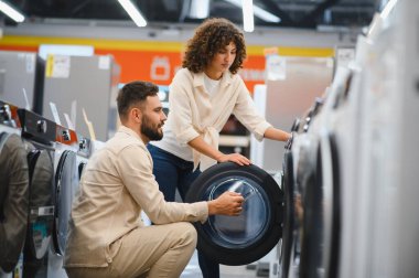 Genç çift, yeni bir çamaşır makinesi seçerken bir yandan da seçeneklerle dolu parlak ve modern bir ev gereçleri mağazasına göz atıyorlar.